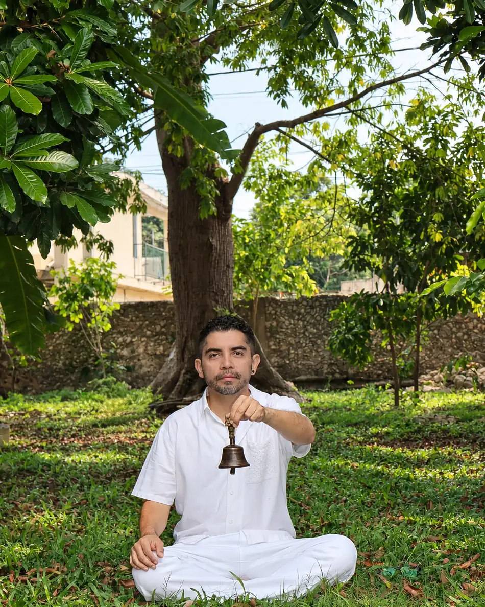 El Dr. Anuar Mena sentado bajo un árbol, con una campana de meditación en la mano.