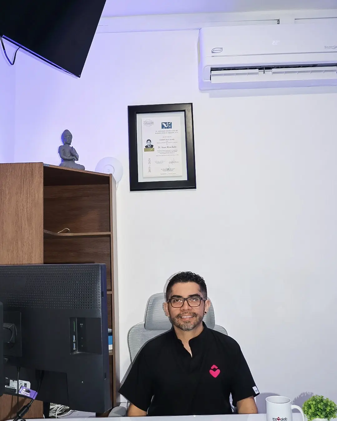 El Dr. Anuar Mena en su consultorio de Inovalab Ticul: los diplomas enmarcados en la pared y un Buda en el librero.