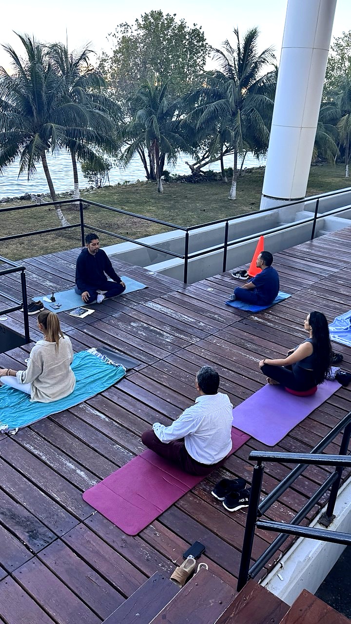 Anuar Mena guiando una práctica de meditación al aire libre, sentado frente a un grupo, con la laguna Nichupté y palmeras al fondo